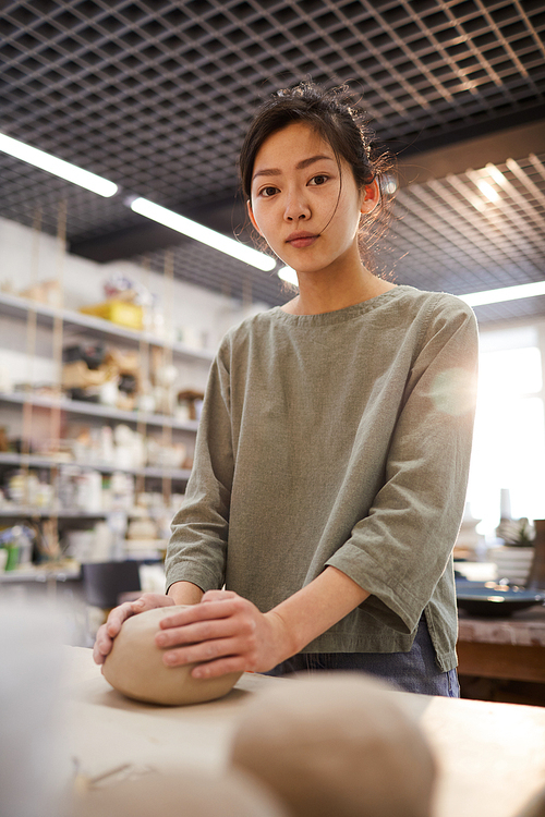 Serious Asian girl in casual clothing standing at desk and working with soft clay in pottery workshop, she 