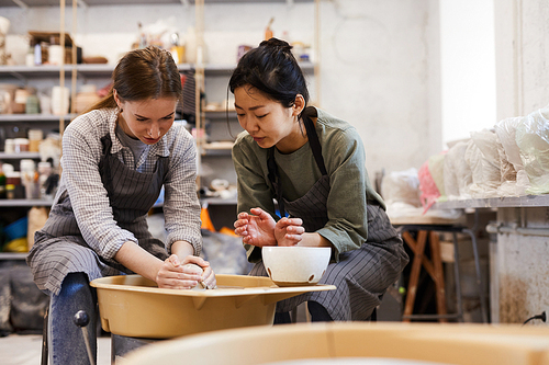 Skilled Asian potter in apron teaching young woman to make clay vase on pottery wheel in modern workshop, pottery course