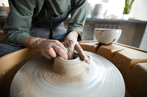 Close-up of unrecognizable woman in dirty hands making hole in clay while modelling vase on pottery wheel