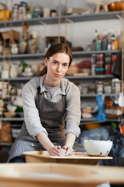 Serious displeased young woman with ponytail forming vase on pottery wheel and  in workshop