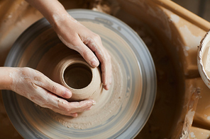 High angle view of unrecognizable female potter with dirty hands sitting at pottery wheel and working with soft clay in workshop