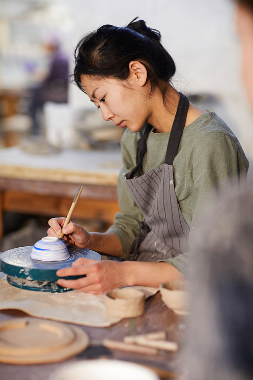 Serious concentrated young Asian woman in apron sitting at table and painting ceramic bowl with paintbrush in workshop