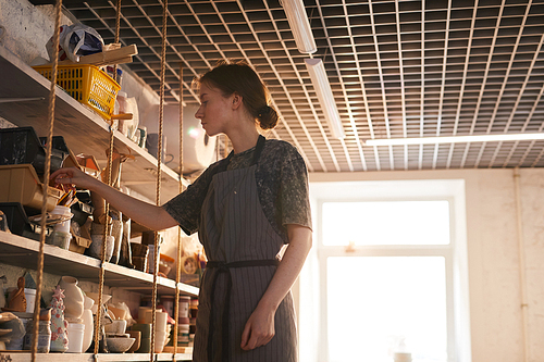 Serious beautiful young lady in gray stripped apron standing at shelves in ceramic studio and choosing tools for work
