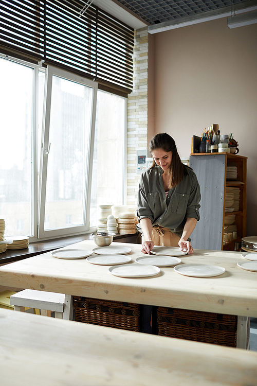 Portrait of modern female artisan arranging handmade ceramic plates in pottery workshop, copy space