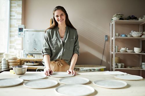 Portrait of smiling female artisan arranging handmade ceramic plates in pottery workshop, copy space