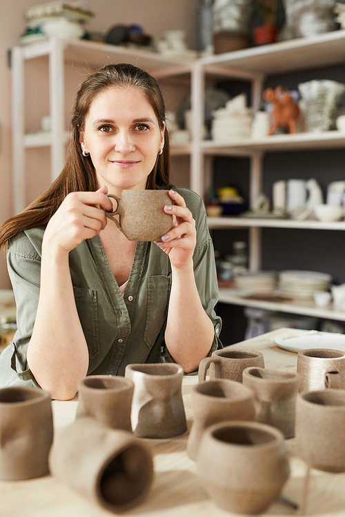 Portrait of smiling female artisan holding handmade ceramic mug while posing in traditional pottery shop