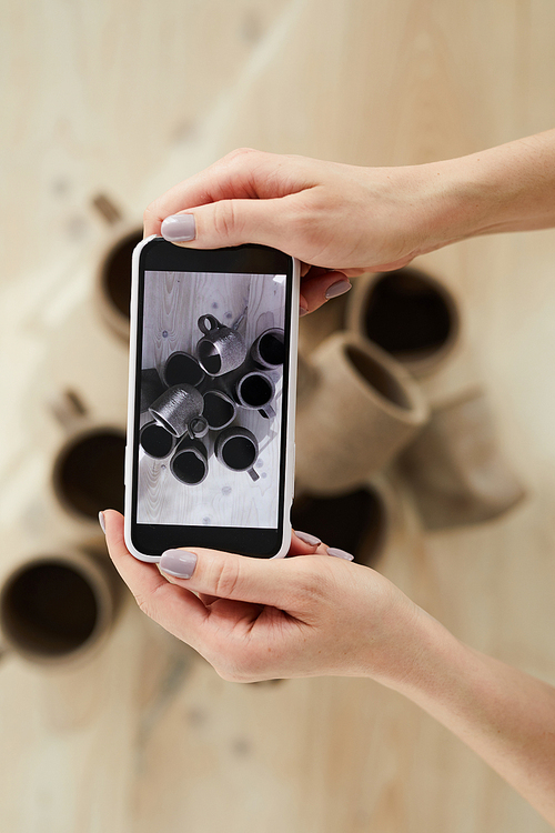 Closeup of female artisan taking smartphone pictures of handmade ceramic items in pottery shop, copy space