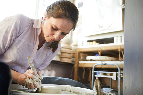 Portrait of female potter creating handmade crockery using wheel in studio, copy space