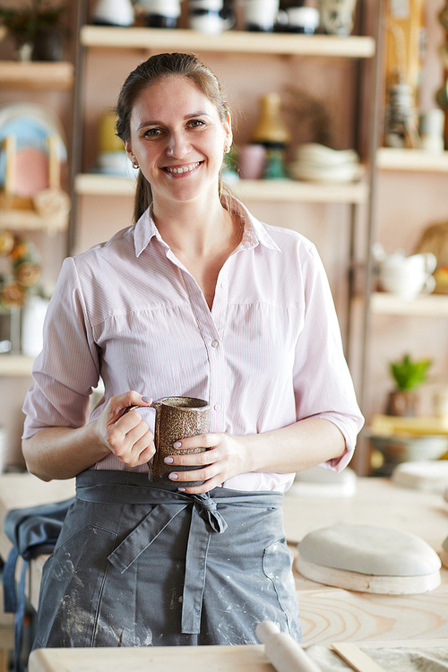 Waist up portrait of cheerful female artisan posing in pottery studio holding handmade mug, copy space
