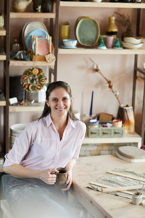 Portrait of cheerful female artisan posing in pottery studio holding handmade mug, copy space