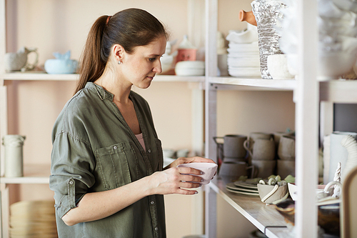 Side view portrait of contemporary female potter putting handmade bowl on shelf in studio, copy space