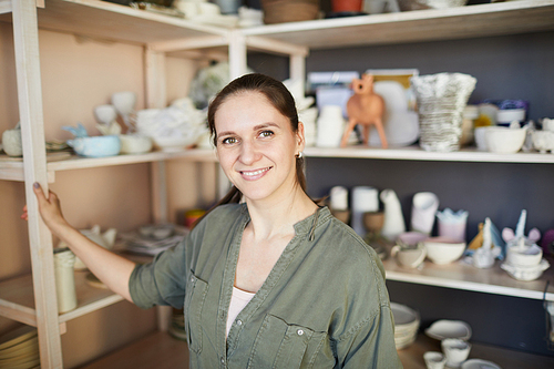 Portrait of smiling female artisan posing in pottery studio against shelves with handmade ceramics, copy space