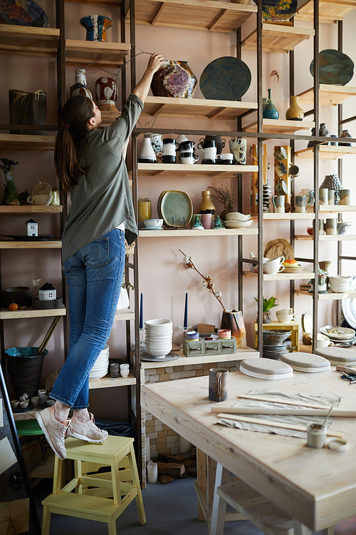 Full length portrait of young woman standing on stool reaching for upper shelf in storage, copy space