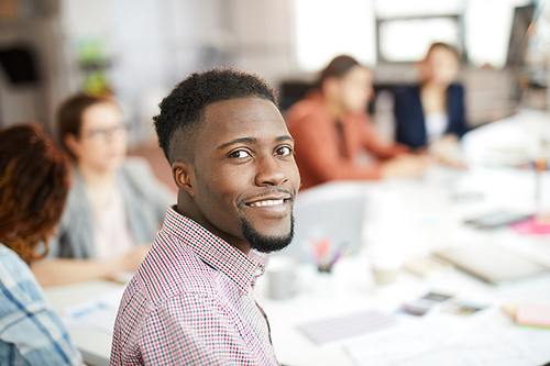 Head and shoulders portrait of young African-American man smiling at camera while sitting at table in business meeting, copy space