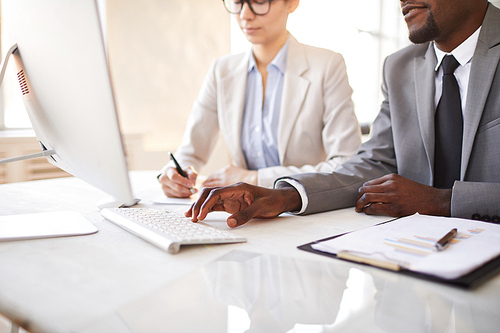 Young African-american broker pressing key on keypad while looking through data on computer screen with colleague sitting next to him