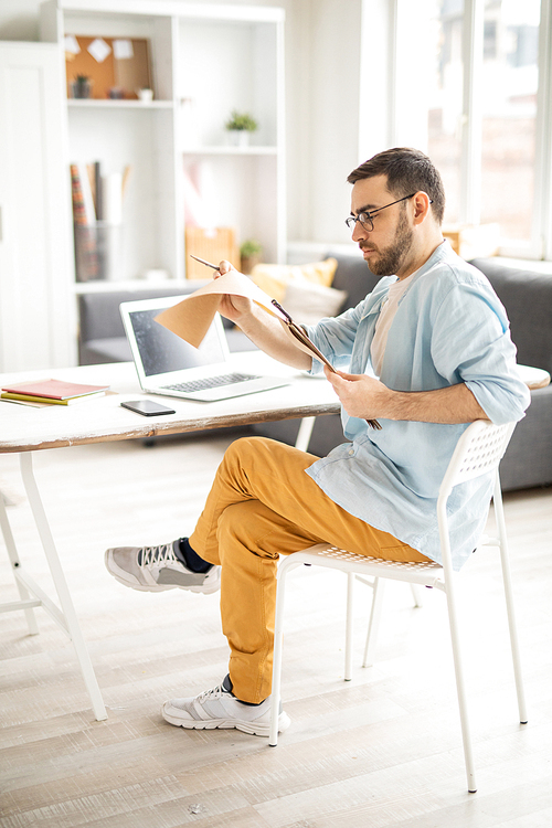 Serious young businessman in casualwear reading his working notes in notepad while sitting by desk in office