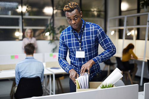 Serious frowning young black man with badge on neck discharged from place of employment checking paper files in box while leaving office