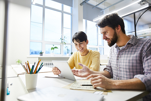 Positive excited young colleagues in casual clothing sitting at table and discussing internet project while using modern tablet in office