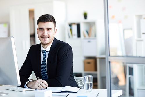 Happy young businessman in formalwear looking at you with smile while sitting by desk in front of computer monitor