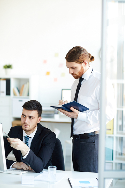 Young confident broker in suit pointing at online data on computer screen while his colleague making notes