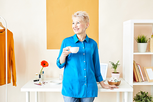 Middle aged laughing woman in smart casual standing by desk in studio while having cup of tea or coffee at break