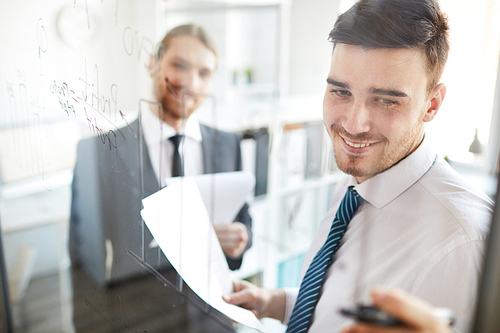 Young cheerful broker in formalwear pointing at data on board while analyzing and explaining it