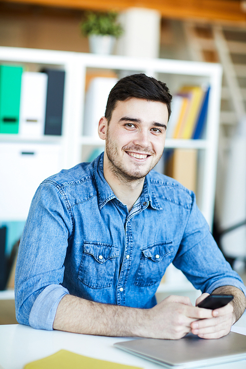 Young cheerful casual manager with smartphone looking at you while sitting by desk in office