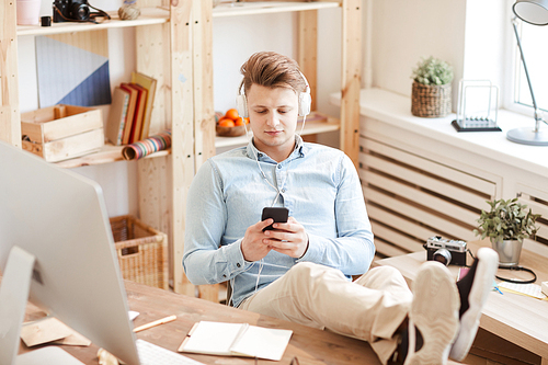 Content relaxed young photographer in wired headphones sitting at table and keeping feet on table while using smartphone in design studio