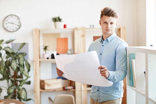 Serious confident young male architect in smart casual outfit standing in modern office and  while analyzing blueprint