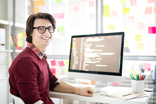 Cheerful young man in casualwear sitting by desk in front of computer monitor during working day in office