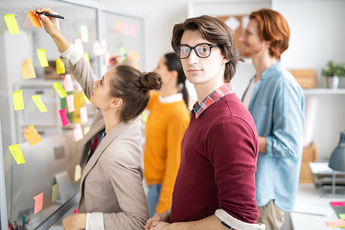 Young serious casual businessman looking at you while standing by noticeboard on background of his colleagues