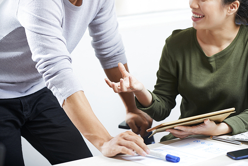 Close-up of positive young lady in green sweater gesturing hand and using tablet while presenting online design sketch to colleague