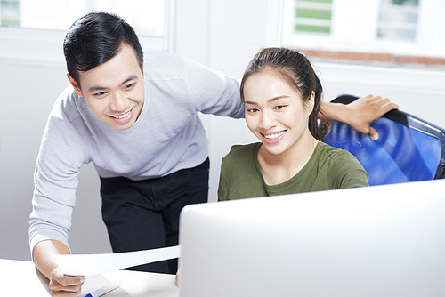 Cheerful confident young Asian computer designers satisfied with results of research looking at monitor while testing app