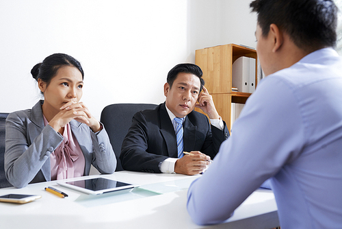 Serious Asian male and female employers in formalwear sitting by desk and listening attentively to young applicant in front of them