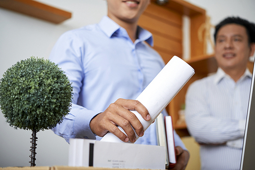 Young new employee holding rolled paper while taking it out of box with his supplies by workplace