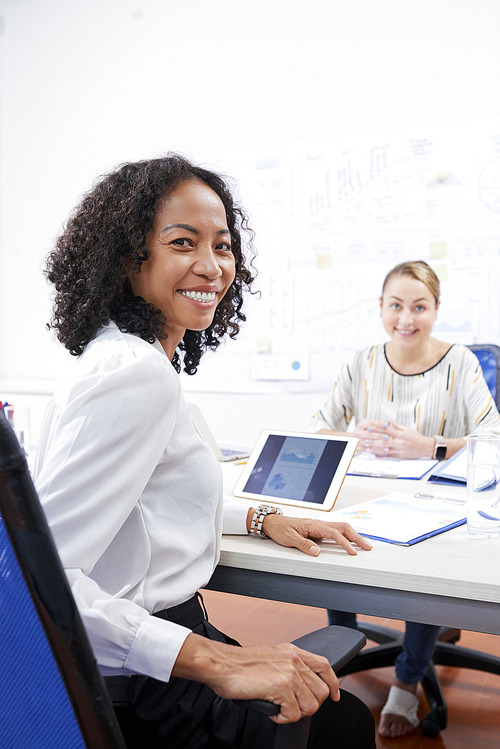 Happy Vietnamese businesswoman sitting at table with female coworker and turning back to look at camera