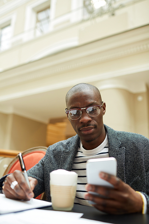 Portrait of contemporary African-American man studying sitting at table in cafe and using smartphone, copy space