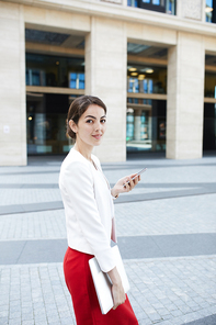 Portrait of beautiful young woman holding smartphone and  while walking outdoors in business area, copy space