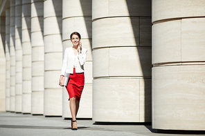 Full length portrait of elegant young woman wearing red skirt walking towards camera along row of pillars and speaking by phone, copy space