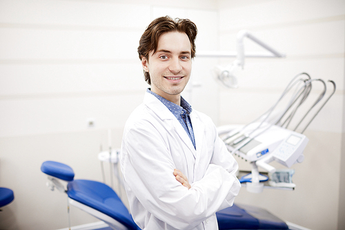 Waist up portrait of smiling young dentist posing in office next to dental chair, copy space