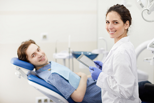 Waist up portrait of female dentist smiling happily at camera while consulting patient in background, copy space