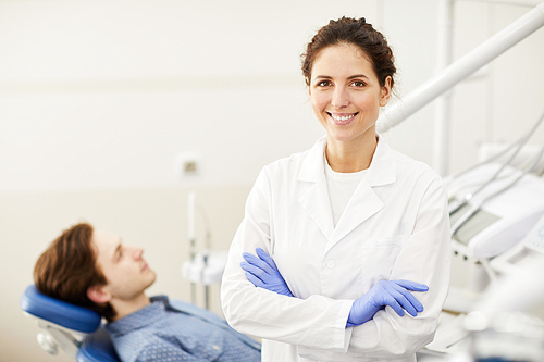 Waist up portrait of female dentist smiling at camera while consulting patient in background, copy space