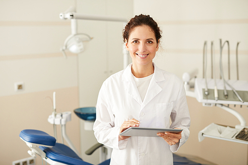 Waist up portrait of confident female dentist smiling at camera holding tablet with dental chair in background, copy space