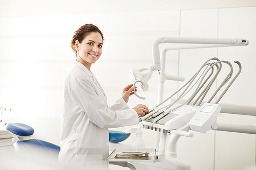 Waist up portrait of confident female dentist smiling at camera while adjusting dental chair , copy space