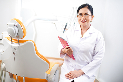 Smiling experienced dentist with tablet computer sitting near dental chair in her office