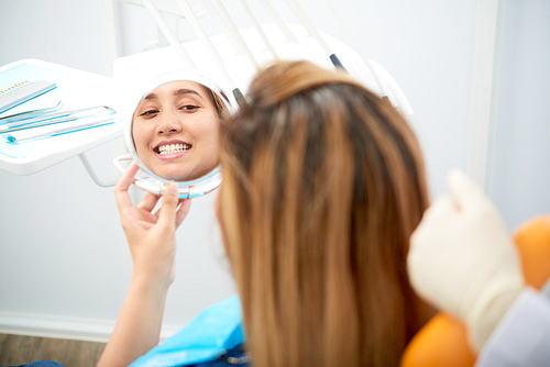 Pretty smiling young Asian woman looking at mirror after whitening teeth at dentistry