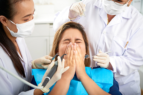 Young frightened woman squinting and covering mouth with hands when dentists trying to treat her teeth