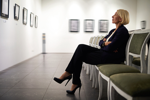 Side view full length portrait of elegant woman sitting on velvet chair looking at pictures in art gallery or museum, copy space