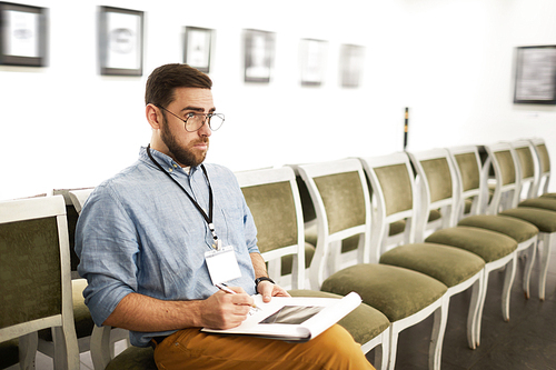 Portrait of bearded young man taking notes while looking at pictures in art gallery or museum, copy space