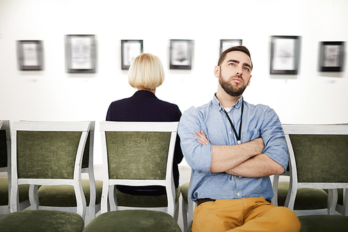 Portrait of bearded young man looking at pictures in art gallery or museum while sitting on velvet chair, copy space